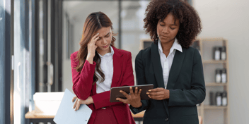 one woman holding tablet displaying profits and loss due to IT downtime while team leader looks on with frustration